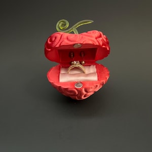 Könnte beinhalten: Eine rote, herzförmige Ringbox mit einem grünen Stiel und einem goldenen Ring im Inneren. Die Box ist geöffnet und zeigt einen goldenen Ring mit einem Diamanten. Die Box befindet sich auf einer dunkelgrauen Oberfläche.