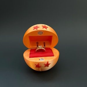 May include: An orange ring box shaped like a dragon ball with red stars. The box is open and a gold ring with a diamond is inside.