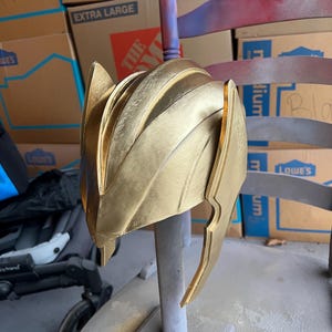 Op de afbeelding: Een gouden helm met een puntige bovenkant en een lang, gebogen stuk dat aan de zijkant uitsteekt. De helm staat op een standaard en lijkt van plastic te zijn.
