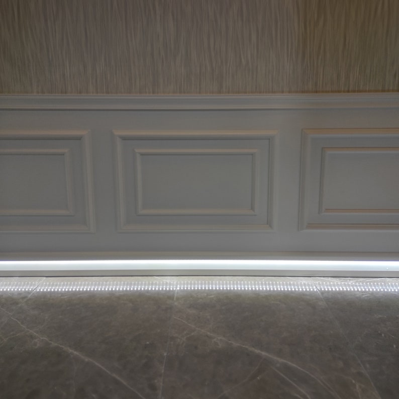 May include: A white wainscoting wall with three panels and a white trim. A thin strip of LED lights is installed along the bottom of the wall, illuminating a gray marble floor.