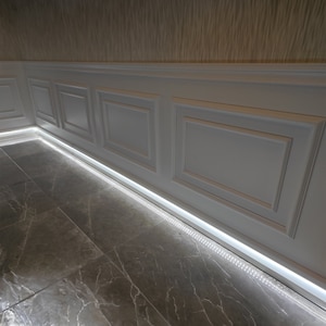 May include: White wainscoting with a decorative molding pattern, installed along a wall with a light brown wallpaper. A strip of LED lights is installed along the baseboard, illuminating the gray marble floor.