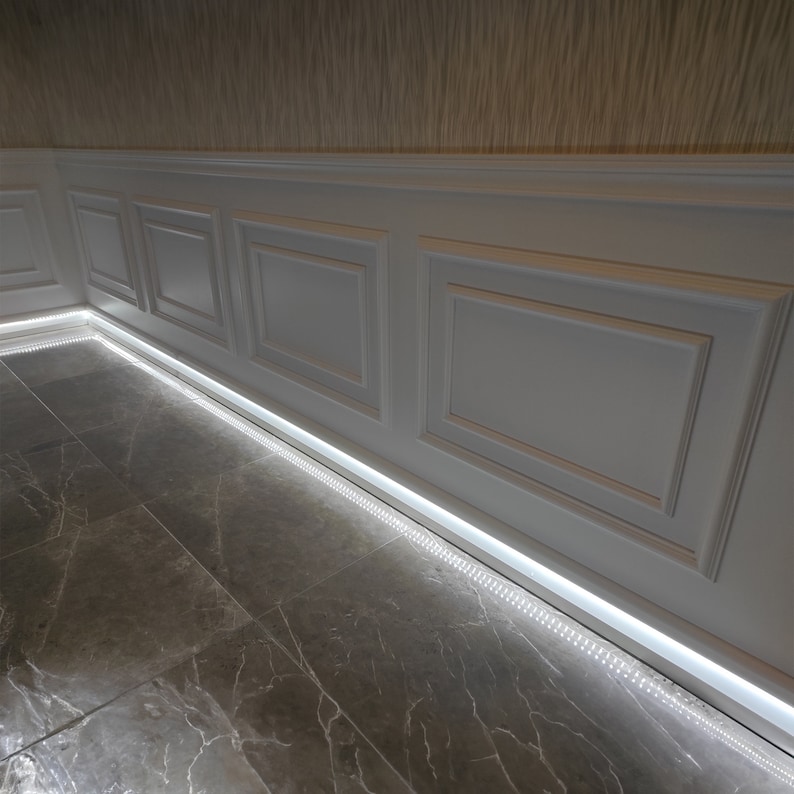 May include: White wainscoting with a decorative molding pattern, installed along a wall with a light brown wallpaper. A strip of LED lights is installed along the baseboard, illuminating the gray marble floor.