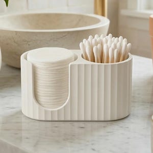 Puede incluir: Un organizador de baño color crema con dos compartimentos. Uno contiene una pila de discos de algodón, el otro bastoncillos de algodón. El organizador tiene un diseño acanalado y se encuentra sobre una encimera de mármol, junto a un lavabo y una planta en maceta.
