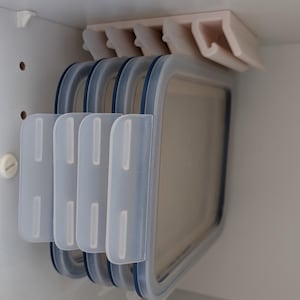 以下が含まれることがあります： 白いキャビネットの内部。透明な長方形の食品容器をいくつか収納する収納ラックがあります。容器には青い蓋があり、きちんと整理されています。ラックは棚の下に取り付けられています。