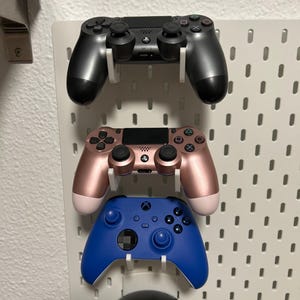 May include: Three game controllers in black, rose gold, and blue are mounted on a white pegboard. The black controller is at the top, followed by a rose gold controller, and a blue controller at the bottom. The controllers are designed for gaming.