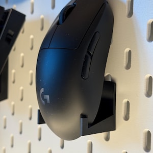 Peut inclure: Une souris d'ordinateur sans fil noire avec une molette de défilement et des boutons latéraux. La souris porte la lettre "G". Elle est montée sur un panneau perforé blanc avec un support noir.