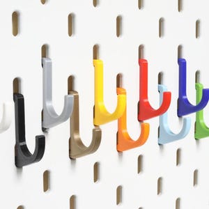 Könnte beinhalten: Eine Reihe farbiger Lochwandhaken in Weiß, Schwarz, Grau, Gold, Gelb, Orange, Rot, Hellblau, Grün und Dunkelgrün. Die Haken sind auf einer weißen Lochwand mit gleichmäßig verteilten Löchern montiert.