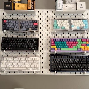 Puede incluir: Una colección de teclados mecánicos exhibidos en una pared perforada blanca. Los teclados varían en color y diseño, incluyendo diseños divididos, de tamaño completo y compactos. Algunos teclados tienen teclas coloridas, mientras que otros son monocromáticos. Estantes arriba sostienen cajas y un cartucho de película.