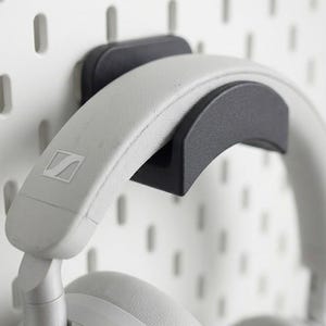 Puede incluir: Unos auriculares blancos cuelgan de un soporte negro para auriculares fijado a una pared perforada blanca. Los auriculares tienen una diadema y almohadillas grises acolchadas. El logotipo de Sennheiser es visible.