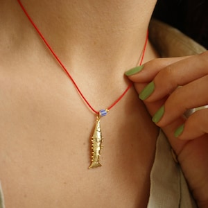 Könnte beinhalten: Eine goldene Fisch-Anhänger-Kette mit einer roten Schnur und einer blau-weiß gestreiften Perle. Die Kette wird von einer Person mit grünem Nagellack getragen.
