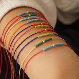 Può includere: Un mucchio di braccialetti colorati in corda con piccole perline e dettagli dorati. I braccialetti sono sovrapposti sul braccio di una persona.