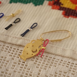 Puede incluir: Encanto de metal dorado en forma de jarrón con una luna creciente y estrellas recortadas. El encanto está unido a un anillo de metal dorado con una cuerda rosa atada.