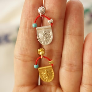 Puede incluir: Pendientes plateados y dorados con un cordón rojo y un acento de cuentas turquesas. Los pendientes presentan un pequeño colgante en forma de escudo con un diseño decorativo. Cierre de poste.