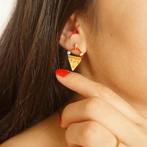 Puede incluir: Pendientes triangulares de oro con una cuenta turquesa y detalles de cuerda roja.