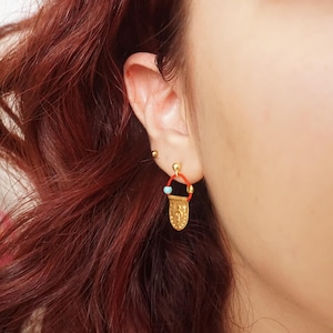 Pode incluir: Brincos dourados com um aro vermelho e uma pequena conta turquesa. Os brincos têm um pendente decorativo em forma de escudo com detalhes em relevo. Os brincos são mostrados numa orelha.