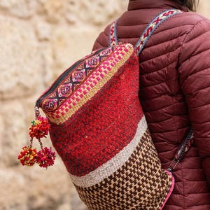 May include: A red and brown woven backpack with a geometric pattern. The backpack has a zippered top, a decorative pom-pom, and patterned shoulder straps. The bag is worn over a burgundy jacket.