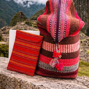 May include: A woven backpack and a matching laptop sleeve featuring horizontal stripes in shades of red, brown, and grey. The backpack has a central zipper with a patterned fabric panel and decorative multi-colored yarn pom-poms attached.