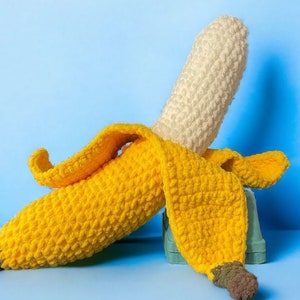Peut inclure: Un jouet en peluche en forme de banane crocheté avec une peau jaune et une pointe blanche. La banane est couchée sur le côté, la peau enroulée.