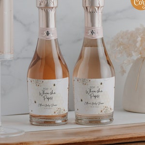 May include: Two mini champagne bottles with pink foil tops and labels that read "Pop It When She Pops!" are displayed with a champagne flute and a vase of white flowers. The bottles are on a wooden tray.