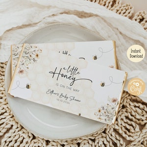 May include: A white and gold baby shower favor with a honeycomb pattern and bees. The text reads "a little honey is on the way" and "Esther's Baby Shower 10.10.2024".