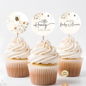 Puede incluir: Tres cupcakes blancos con glaseado blanco y chispas doradas coronados con adornos redondos para cupcakes de baby shower, blancos y dorados. Los adornos presentan una abeja, flores y el texto "a little Honey is on the way" y "Welcome Baby Williams".