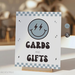 May include: A rectangular sign featuring a light blue and white checkered border at the top and bottom. The center displays a light blue smiley face with lightning bolt eyes, surrounded by small star icons, with the text CARDS AND GIFTS in black.