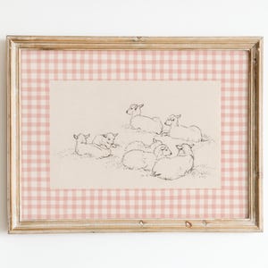May include: A framed print with a pink and white gingham border. The print features a black and white illustration of a group of lambs lying down.