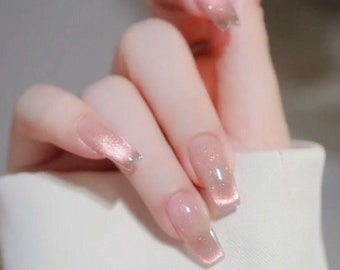 May include: Close-up of a hand with long, square-shaped nails painted with a light pink glitter polish. The nails have a clear, iridescent finish.