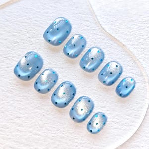 Peut inclure: Un ensemble de faux ongles bleus clairs, de forme ovale et à finition brillante. Chaque ongle est orné de petits points bleu foncé. Les ongles sont présentés sur une surface transparente et présentent un léger scintillement.