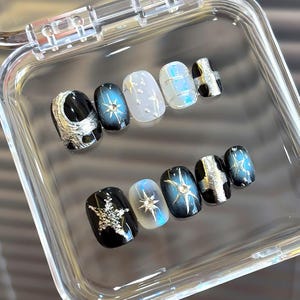 May include: A set of press-on nails in a clear plastic case. The nails feature various designs, including silver moons, stars, and geometric patterns. Colors include black, silver, white, and iridescent blue.