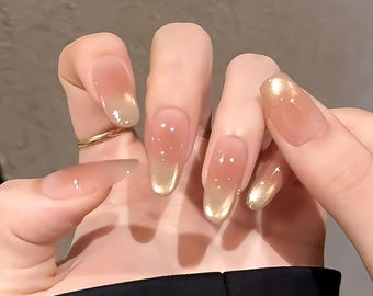 May include: Close-up of a hand with several nude-colored oval-shaped press-on nails with a gold glitter accent at the tip.