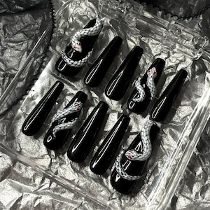 Puede incluir: Juego de uñas postizas negras en forma de ataúd. Algunas uñas presentan un diseño de serpiente blanca con detalles rojos. Las uñas se muestran en un recipiente de plástico transparente, ideal para los amantes del arte de uñas.