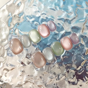 Puede incluir: Una colección de diez uñas postizas ovaladas en varios colores pastel, incluyendo rosa, verde y lavanda. Las uñas tienen un acabado brillante y un efecto ojo de gato, creando una apariencia brillante. Están dispuestas sobre una superficie texturizada y reflectante.