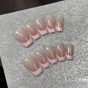 Könnte beinhalten: Ein Set künstlicher Nägel mit klassischem French-Maniküre-Design. Die Nägel haben eine hellrosa Basis, eine nude-farbene Spitze und eine weiße Spitze. Die Nägel sind auf einer silbernen Oberfläche angeordnet.