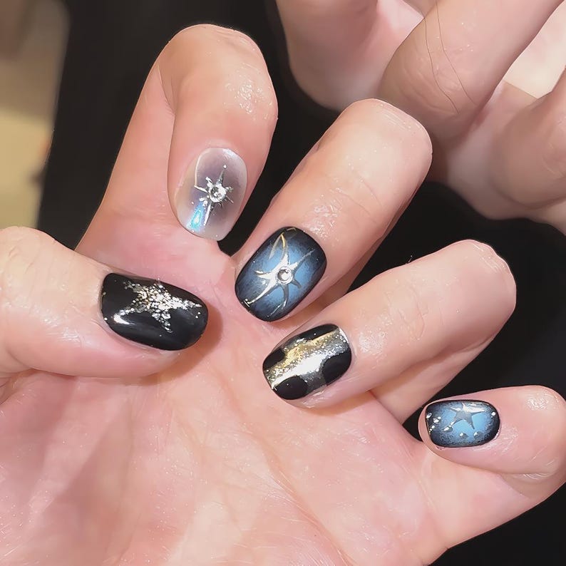 May include: A hand with manicured nails featuring a celestial theme. The nails are painted in shades of black, blue, and silver, with star and galaxy designs. Some nails have a gradient effect, and others feature glitter accents.