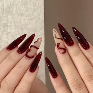 Pode incluir: Unhas longas e pontiagudas em forma de estilete pintadas de vermelho escuro com um design de cobra em duas das unhas. O design da cobra é feito em um tom de vermelho mais claro.