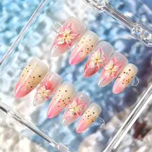 Puede incluir: Juego de diez uñas artificiales con diseño floral. Cada uña presenta un degradado de rosa y beige, adornada con una flor blanca y rosa y pequeños puntos marrones. Las uñas se exhiben en un recipiente de plástico transparente.