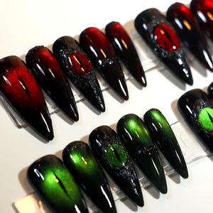 May include: A selection of long, pointed false nails in various designs. Some nails are black with red or green cat-eye effects, while others feature textured accents and a central eye design. The nails are arranged on a clear surface.