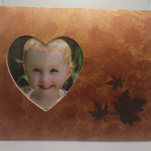 Könnte beinhalten: Ein herzförmiger Bilderrahmen mit einem Ausschnitt in der Mitte, der ein Foto eines lächelnden Kindes zeigt. Der Rahmen ist aus Holz und in einem warmen, rostigen Orangeton lackiert. Der Hintergrund zeigt ein Design aus stilisierten Blättern in einem dunkleren Braunton.