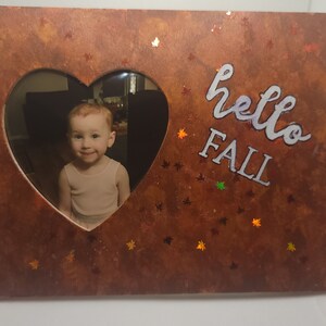 May include: A heart-shaped frame with a photo of a young child inside. The frame is made of wood and has a brown, rustic finish. The text "hello fall" is written in white paint on the frame.