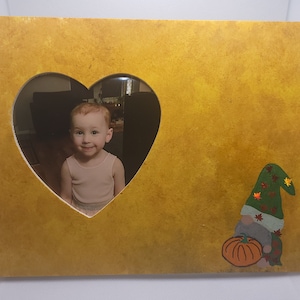 Könnte beinhalten: Ein gelber Holzbildrahmen mit einem herzförmigen Ausschnitt. Der Ausschnitt zeigt ein Foto eines kleinen Kindes. Ein kleiner Gnom mit grünem Hut und einem Kürbis ist auf den Rahmen gemalt.