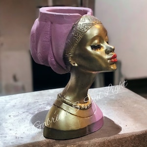 May include: Gold-coloured planter shaped like a woman's head with a pink turban. The planter has a gold necklace and red lips.