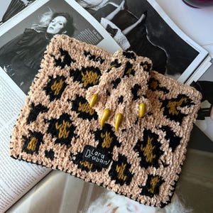 Puede incluir: Un bolso de mano con estampado de leopardo beige y negro con un asa a juego. El bolso tiene un aspecto suave y texturizado y presenta una etiqueta que dice "Lara Drabain". El bolso de mano está adornado con adornos amarillos en forma de garra.