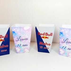 Peut inclure: Quatre contenants en papier blancs de forme trapézoïdale. Deux présentent le logo Red Bull et le nom "Maho" avec "15 ans". Les deux autres affichent le nom "Louna" et "11 ans" sur un fond pastel.