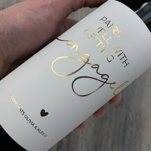 Peut inclure: Bouteille de vin blanc avec une étiquette en feuille d'or qui dit "Pairs well with getting engaged" et "Congrats Olivia & Alexander".