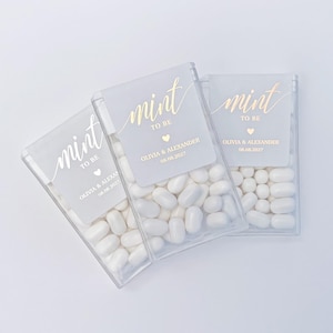 May include: Three clear plastic boxes filled with white mints. Each box has a label with the words "mint to be" in gold script, a heart, and the names Olivia & Alexander with the date 08.08.2027.