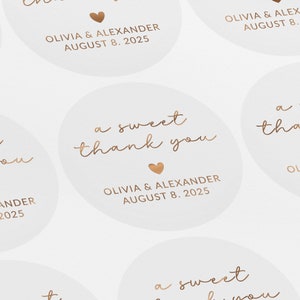 May include: A set of round, white stickers with gold lettering. The stickers say "a sweet thank you" with a small heart and "Olivia & Alexander August 8, 2025".