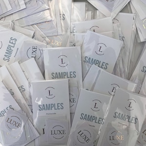 May include: An overhead shot of a pile of white sample packets and stickers. The packets are labeled "SAMPLES" in a gray font, with a stylized "L" logo. Some stickers read "LUXE".