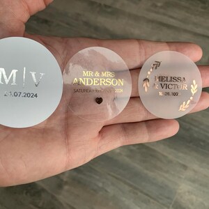 May include: Three round wedding favor stickers. One white sticker with silver lettering says "M/V 24.07.2024". One clear sticker with gold lettering says "MR & MRS ANDERSON SATURDAY 10TH MAY 2024". One clear sticker with gold lettering and a floral design says "MELISSA & VICTOR 10.08.2024".