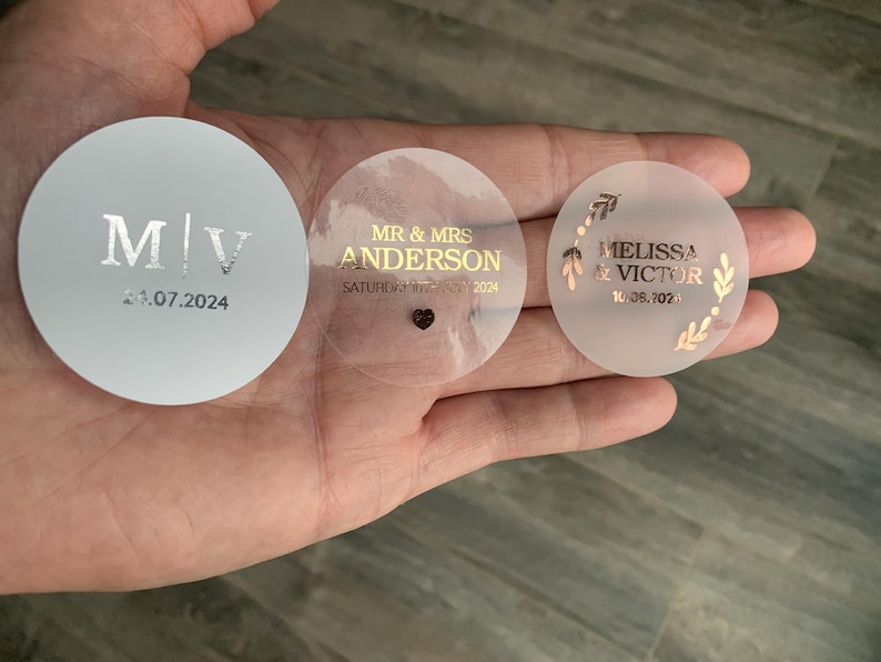 May include: Three round wedding favor stickers. One white sticker with silver lettering says "M/V 24.07.2024". One clear sticker with gold lettering says "MR & MRS ANDERSON SATURDAY 10TH MAY 2024". One clear sticker with gold lettering and a floral design says "MELISSA & VICTOR 10.08.2024".
